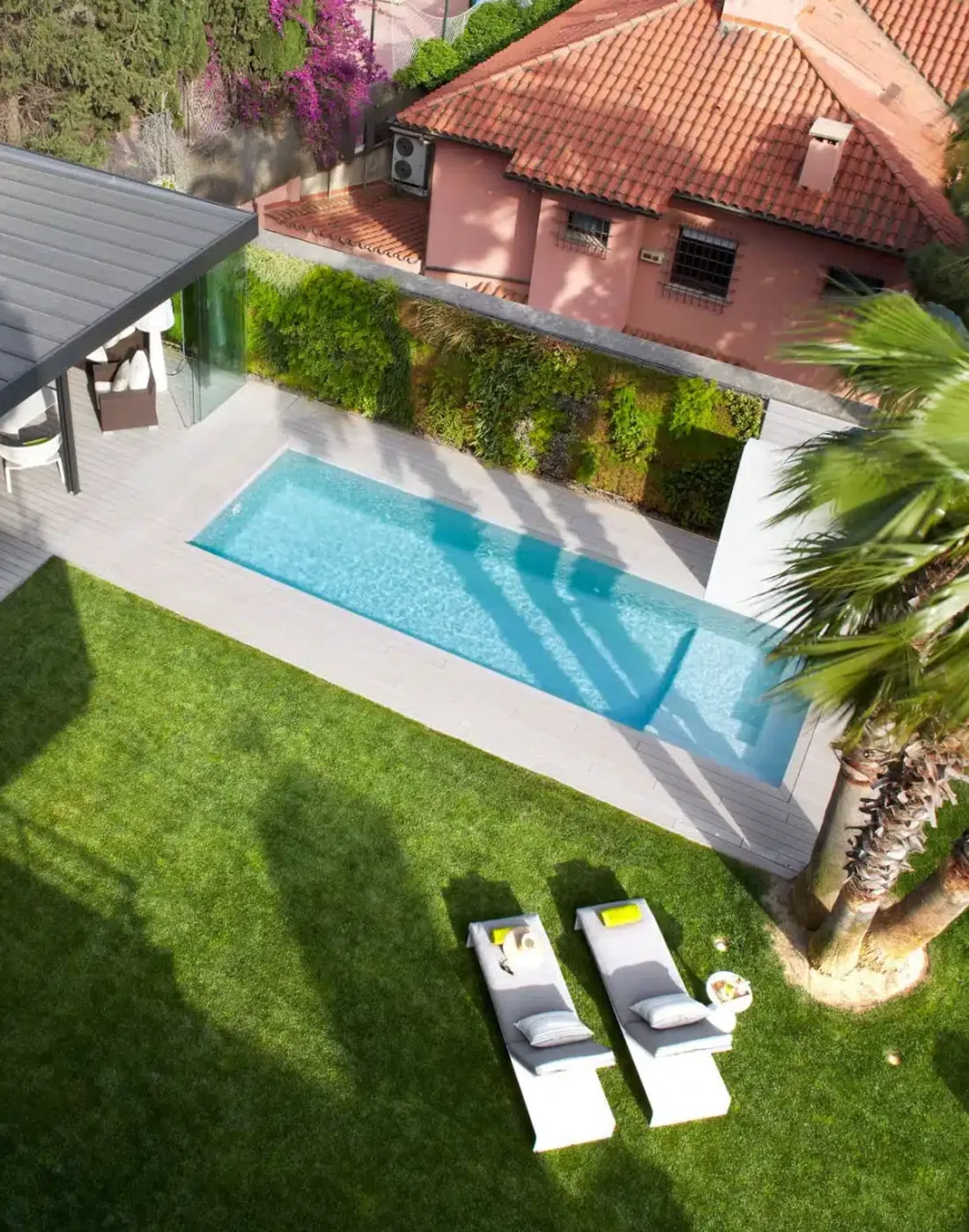 Vista desde arriba de un jardín con césped, jardín vertical, porche acristalado, piscina alargada y dos tumbonas